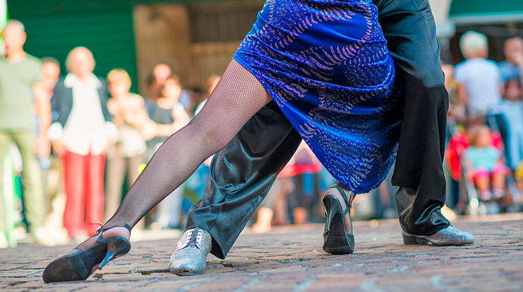 Visão somente das pernas dos bailarinos dançando tango, sendo que em primeiro plano está a mulher de vestido azul, meia calça e sapato e, atrás dela, o homem de calça preta e sapatos claros.