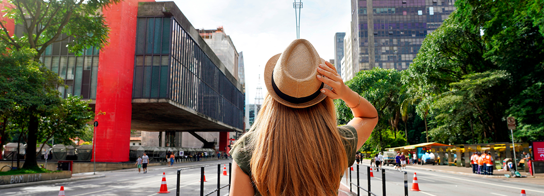 Avenida Paulista com o Masp à esquerda e o Parque Trianon à direita. No centro, em primeiro plano, uma mulher de costas com chapéu panamá.