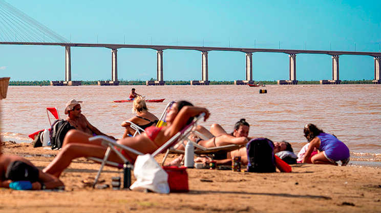 Pessoas em cadeiras de praia no primeiro plano, ao fundo, a ponte de Rosário.