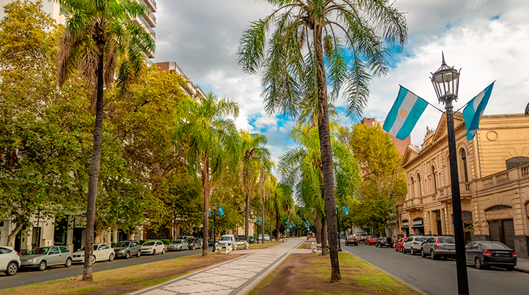 Rua arborizada com carros estacionados e bandeira argentina pendurada em poste.