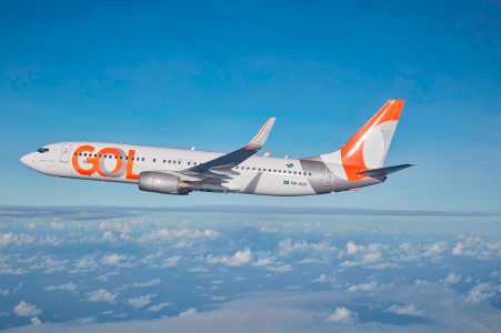 Imagem de uma avião branco, com o logo da empresa GOL e detalhes do logotipo nos estabilizadores e leme traseiros; o avião está sobrevoando as núvens, direcionado para a esquerda