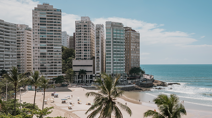 Vista panorâmica de uma praia do Guarujá, com prédios em torno da praia, coqueiros, faixa de areia clara e mar que se estende por todo o lado direito da imagem.