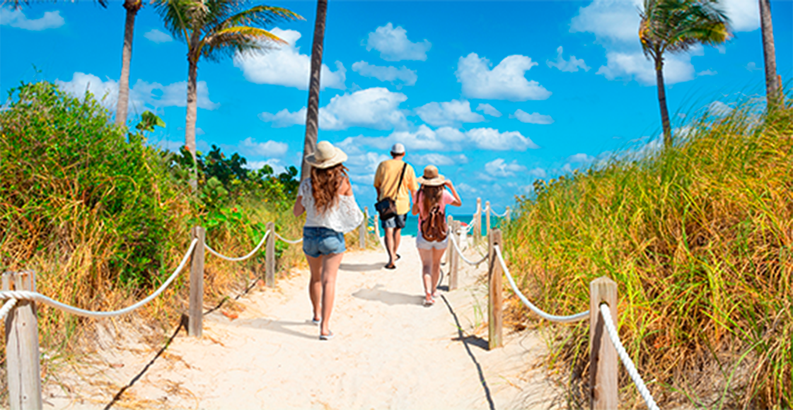 Três pessoas de costas adentram uma praia de Miami por um caminho com cercas.