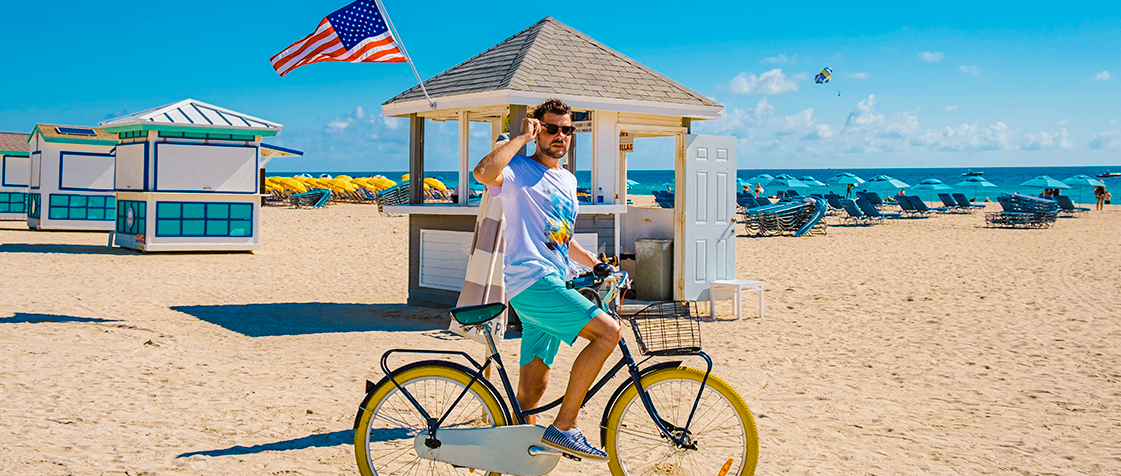 Homem montado em uma bicicleta na praia de Miami, com cadeiras, guarda-sóis e quiosques ao longo da faixa de areia.