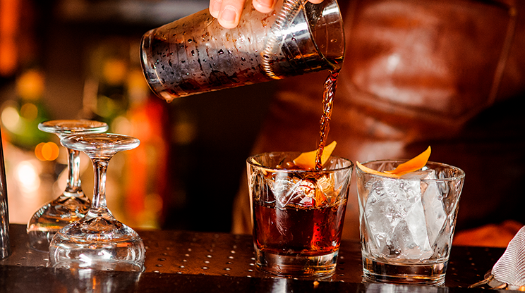 Balcão de bar na Vila Madalena em primeiro plano, com taças viradas para baixo e a mão de um bartender dividindo um drink da coqueteleira em dois copos com gelo.