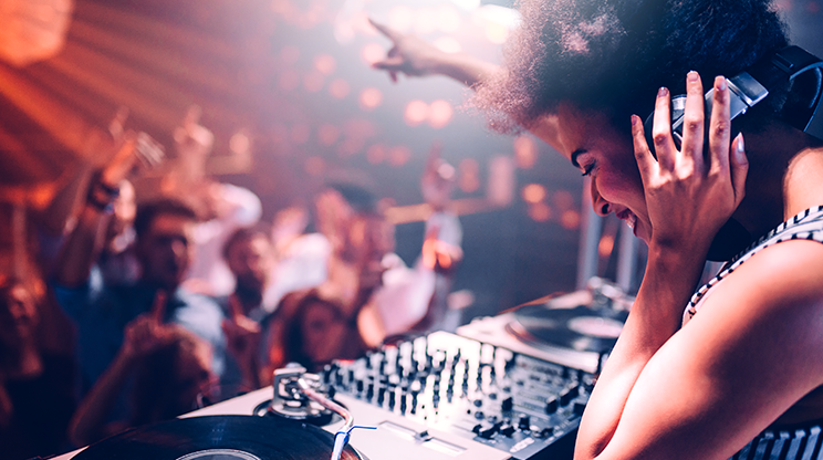 Festa de música eletrônica com uma DJ em primeiro plano, segurando o fone com uma das mãos e apontando para o público com a outra. Na frente dela, está o equipamento para discotecar e o público curtindo a festa.