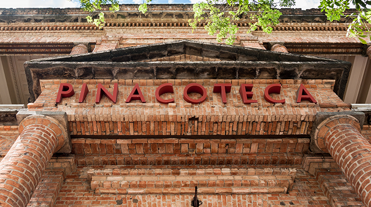 Fachada do museu em tijolos e com o nome "Pinacoteca" em letras grandes.