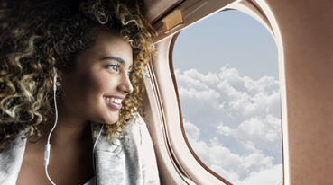 Young mixed race woman smiles while looking through window on aircraft. She is wearing earbuds.