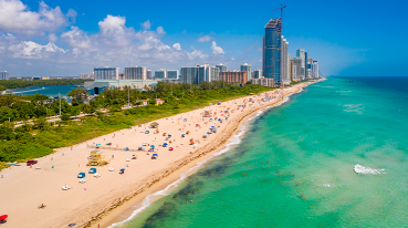Praia de Haulover Park, em Miami, com uma faixa de areia clara que se estende em direção ao mar azul-turquesa. Na areia, temos guarda-sóis e cadeiras, e a praia é cercada de vegetação e edifícios.