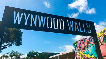 Fachada da entrada da galeria a céu aberto Wynwood Walls, com o nome gravado em uma placa grande e um pequeno pedaço de parede grafitado.