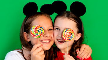 Duas meninas entre 5 e 8 anos abraçadas, com orelhinhas de Mickey, cada uma cobrindo um dos olhos com um pirulito colorido.
