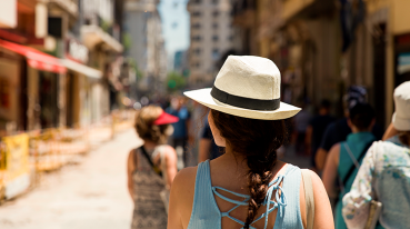 Mulher de costas, com blusa azul, cabelos trançados e chapéu panamá, andando pelas ruas de Buenos Aires em pleno verão.