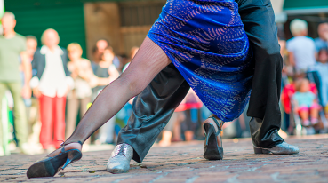 Visão somente das pernas dos bailarinos dançando tango, sendo que em primeiro plano está a mulher de vestido azul, meia calça e sapato e, atrás dela, o homem de calça preta e sapatos claros.